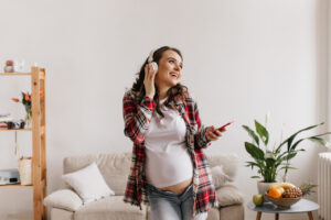 happy brunette pregnant woman in denim pants and white tee listening to music in headphones. cool young mother in jeans holds phone and dance in living room.