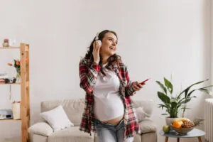 happy brunette pregnant woman in denim pants and white tee listening to music in headphones. cool young mother in jeans holds phone and dance in living room.