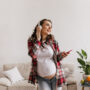 happy brunette pregnant woman in denim pants and white tee listening to music in headphones. cool young mother in jeans holds phone and dance in living room.