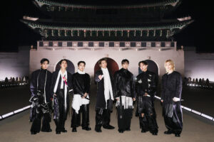 "V, JIN, SUGA, RM, JUNG KOOK, J-HOPE, and JIMIN dressed in fashionable black and white outfits stand outdoors at night in front of an ornate, traditional East Asian building with arches and a large decorative roof."