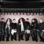 "V, JIN, SUGA, RM, JUNG KOOK, J-HOPE, and JIMIN dressed in fashionable black and white outfits stand outdoors at night in front of an ornate, traditional East Asian building with arches and a large decorative roof."