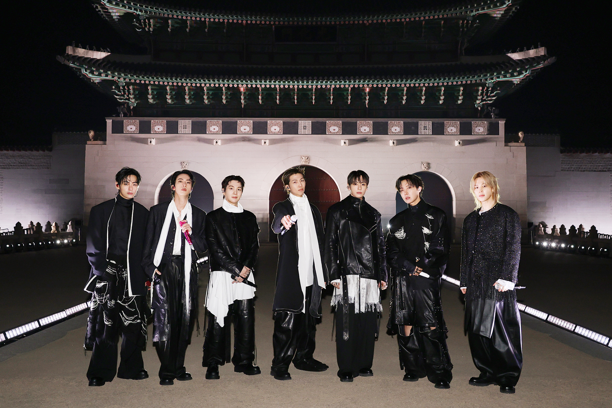 "V, JIN, SUGA, RM, JUNG KOOK, J-HOPE, and JIMIN dressed in fashionable black and white outfits stand outdoors at night in front of an ornate, traditional East Asian building with arches and a large decorative roof."